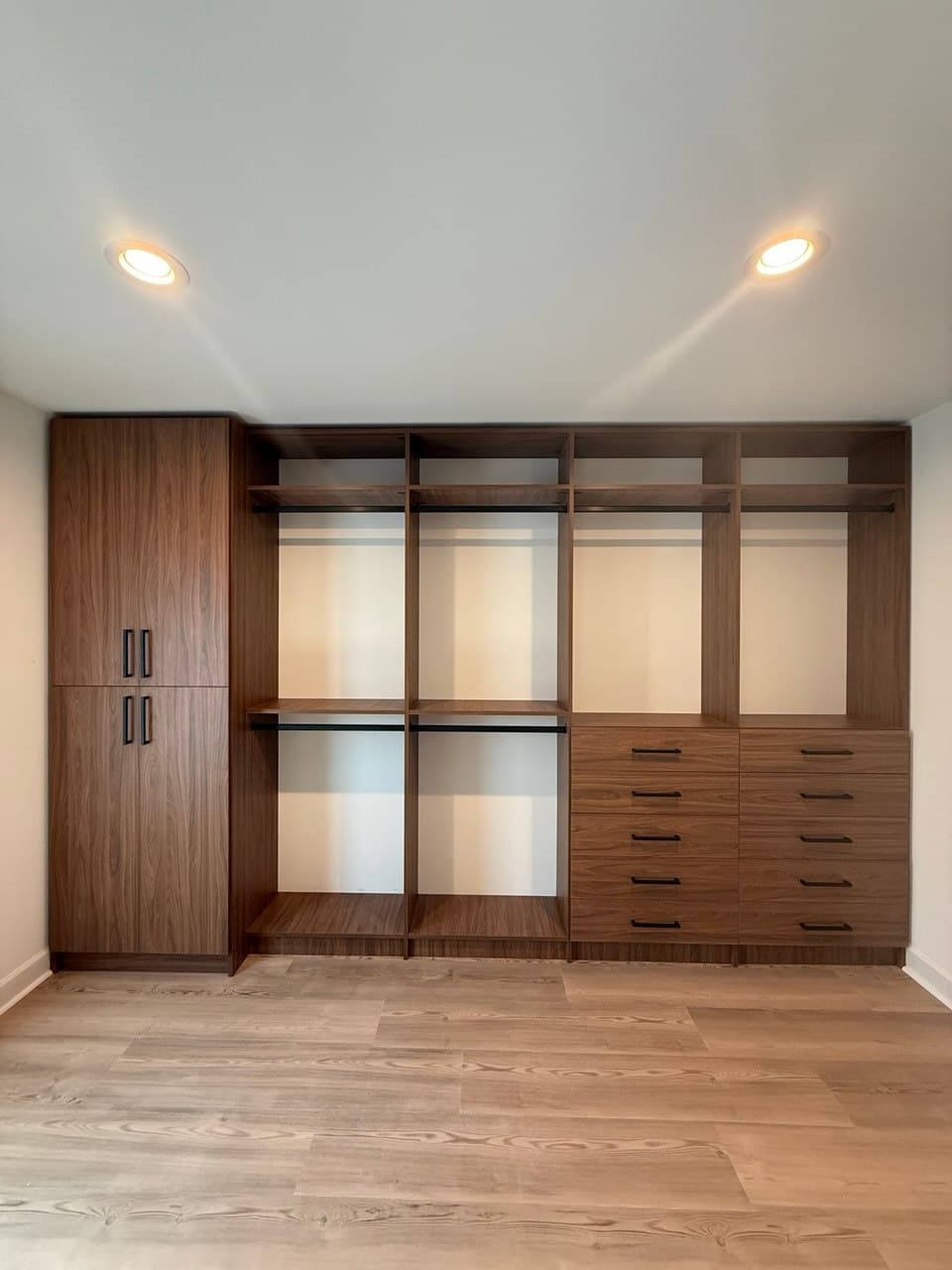 Custom walnut-toned closet system in an empty room: a tall double-door cabinet on the left, four sections of hanging rods and shelves, and a bank of ten drawers on the right.